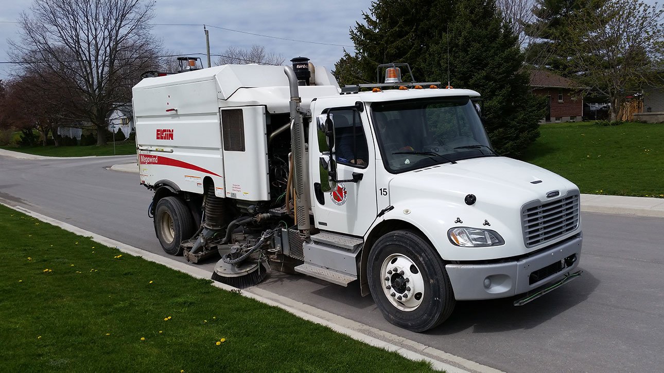 small vehicle with side sweeper attached, sweeping the curb on a residential street